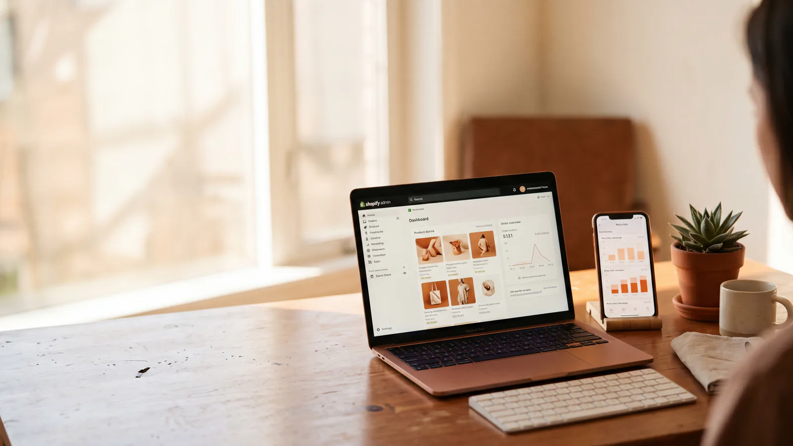 Warm bureautafereel met open laptop die een Shopify-dashboard toont en smartphone met Meta Ads-statistieken, compact toetsenbord en klein cactusje op warm eiken bureau